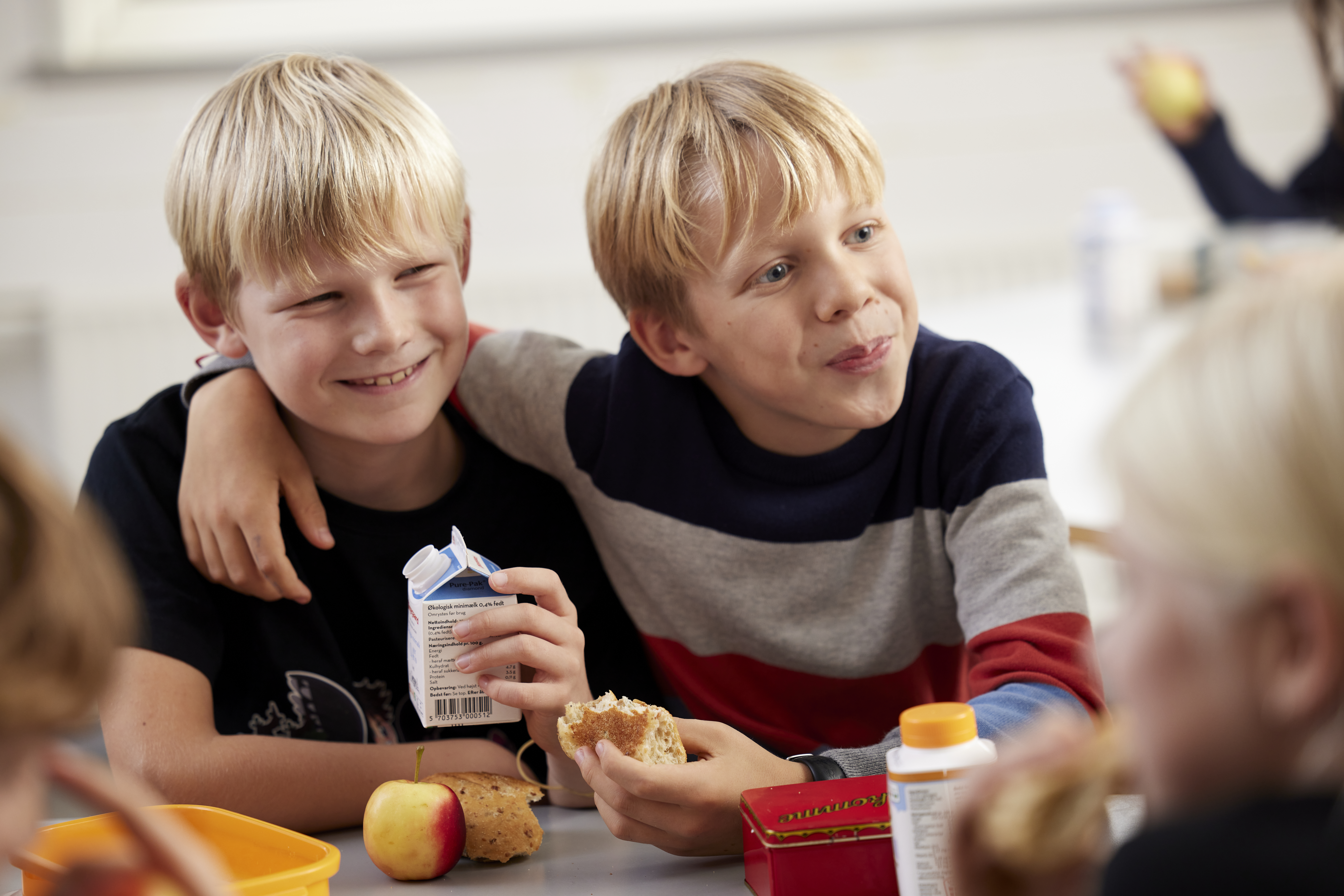 A boy with his arm around another boy at lunch in school (drinking milk)