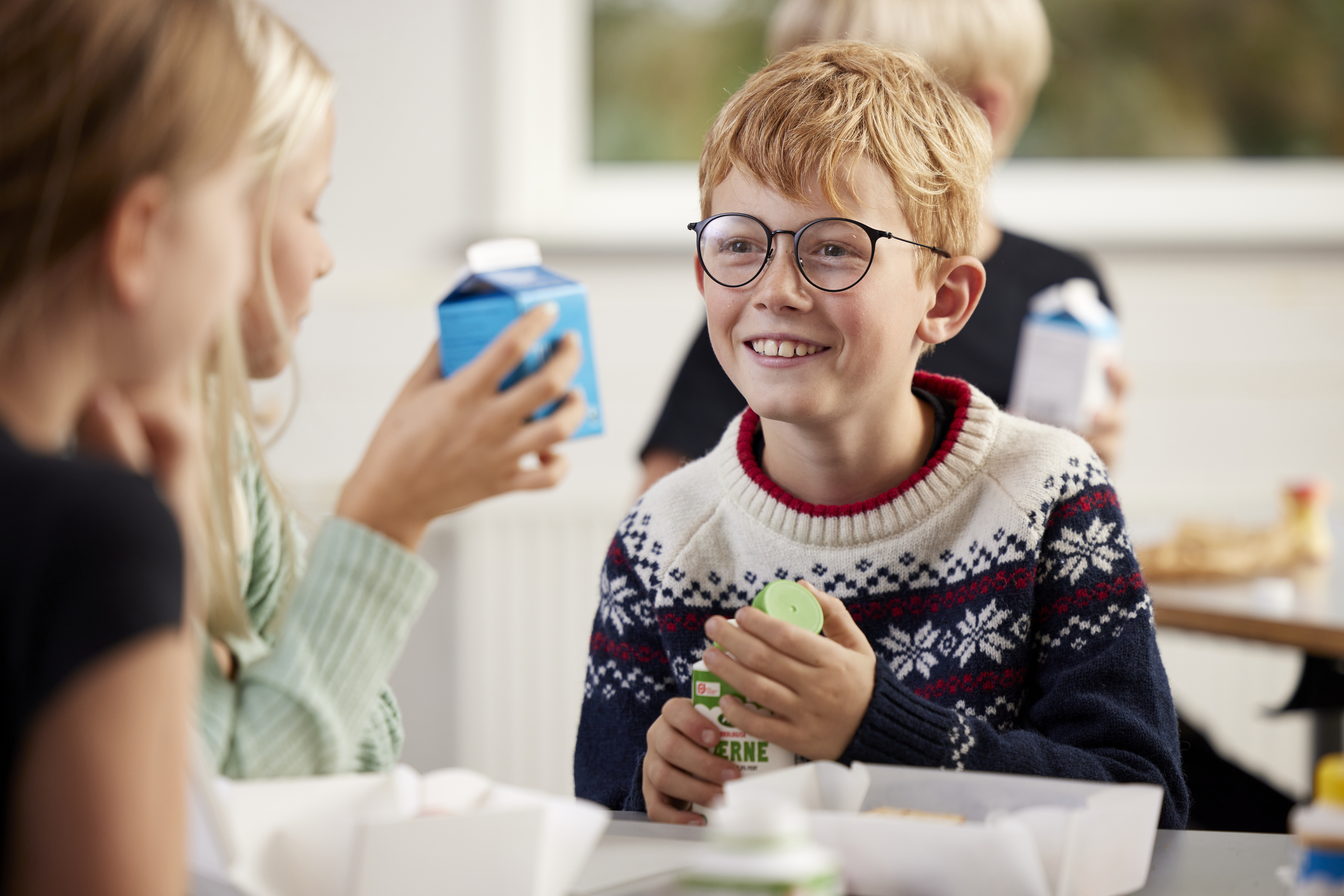 Childen at lunch time at school. A boy with glasses is smiling at a girl holding a school milk carton